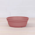 A terracotta-colored plastic Re-Play Bowl from REPLAY sits on a light wooden surface, with a white tiled wall in the background. Made from recycled plastic bowls, this product is designed to catch water draining from plant pots. Its simple, smooth design is practical and understated.