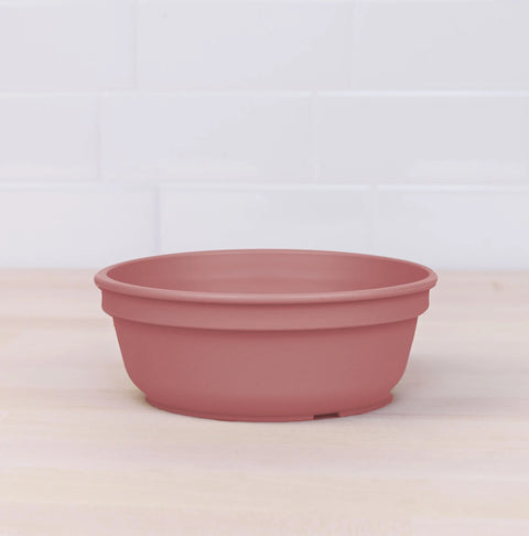 A terracotta-colored plastic Re-Play Bowl from REPLAY sits on a light wooden surface, with a white tiled wall in the background. Made from recycled plastic bowls, this product is designed to catch water draining from plant pots. Its simple, smooth design is practical and understated.