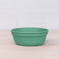 A Re-Play Bowl by REPLAY, crafted from recycled green plastic, is placed on a light wooden surface. The background features a white tiled wall. The bowl boasts a simple design with no additional markings or patterns, ensuring it is safe for kids to use.
