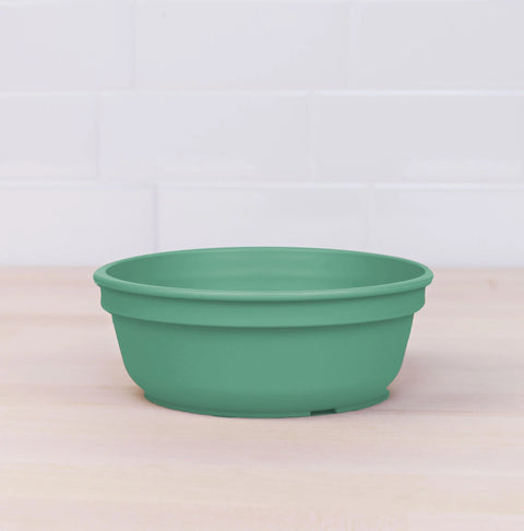 A Re-Play Bowl by REPLAY, crafted from recycled green plastic, is placed on a light wooden surface. The background features a white tiled wall. The bowl boasts a simple design with no additional markings or patterns, ensuring it is safe for kids to use.