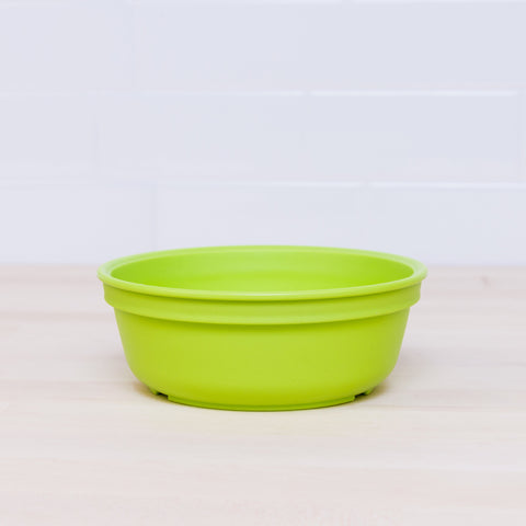 A vibrant Re-Play Bowl from REPLAY, in bright green, sits on a light wooden surface. The background showcases glossy, white tiled walls. Crafted from recycled plastic, this shallow round bowl is also safe for kids.