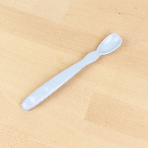 The Re-Play Infant Spoon by REPLAY is a light blue plastic spoon designed for infants. It features a small, rounded bowl and a slightly curved handle with three circular indents, all placed on a wooden surface. This piece of sustainable tableware is perfect for your baby's mealtime.