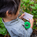 A young child wearing a gray shirt examines a small spiral notebook with pictures and descriptions of insects and plants, part of their Tiger Tribe Bug Spotter Kit by TIGER TRIBE. In the child's other hand is a green magnifying glass. They are standing among green leafy plants in what appears to be an outdoor exploration.