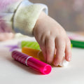 A child's hand reaches for the vibrant Tiger Tribe Junior Markers by TIGER TRIBE on a white surface, adorned in a long-sleeved, pastel-colored shirt. The non-toxic markers are green, yellow, and pink, with some lying horizontally, against a blurred background.