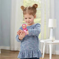 A little girl with light brown hair in two buns is looking at her VTech Paw Patrol Sky Learning Watch. She is wearing a blue sweater with a ruffled hem. In the background, there is a white table with a book and a lamp, and a window with sheer curtains.